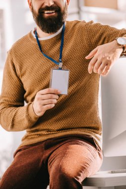 ofis masası üzerinde oturan ve işyerinde boş güvenlik etiketi tutan sakallı yetişkin iş adamı görünümünü kırpılmış