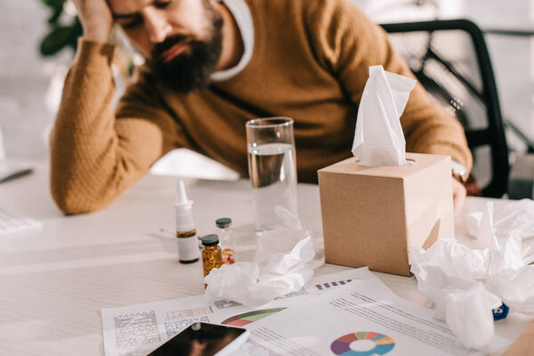selective focus of tired sick businessman sitting at workplace with medicine, pills and tissue box on office desk 