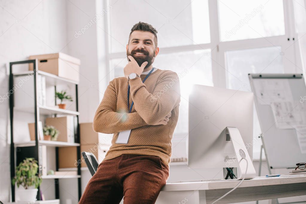 smiling bearded adult businessman with blank security tag touching chin and looking at camera in office