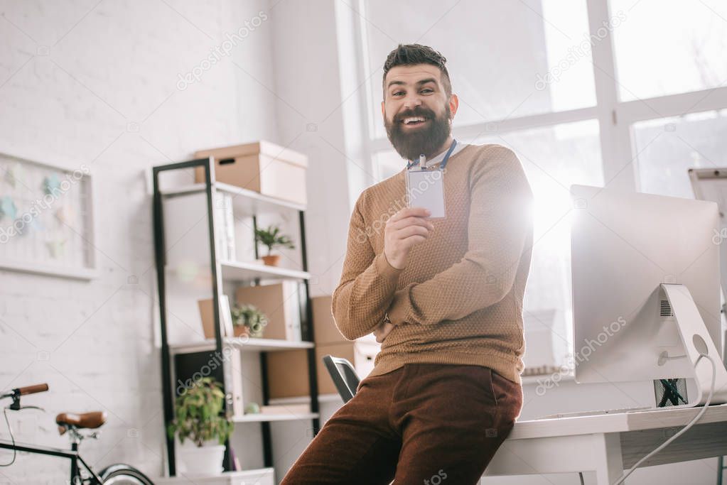 smiling bearded adult businessman sitting on office desk and holding blank security tag at workplace