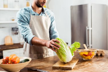 cropped shot of young man in apron holding napa cabbage and cooking in kitchen clipart