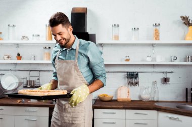 önlük ve taze ev yapımı pizza ile fırın tepsisi elinde tutacak gülümseyen genç adam