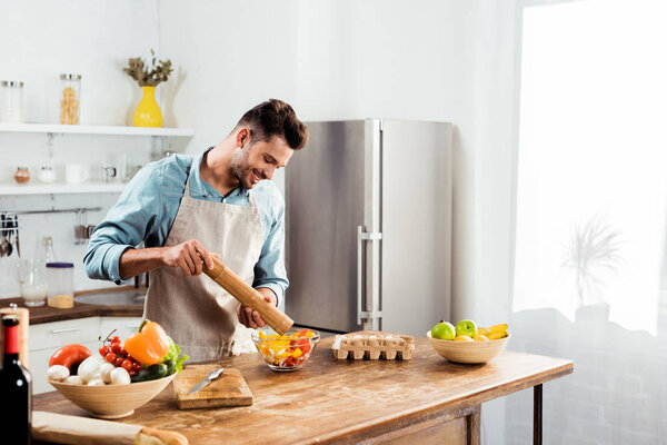 handsome smiling young man adding pepper with mill into salad