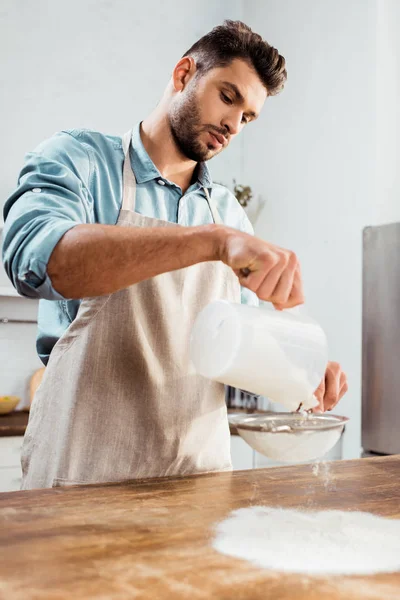 エプロン台所のテーブルに小麦粉をふるいでハンサムな若い男の低角度のビュー  — 無料ストックフォト