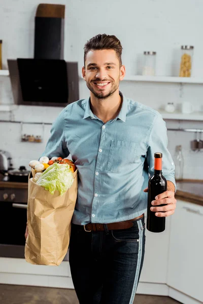 Krásný Usměvavý Mladý Muž Drží Pytel Potravinami Láhev Vína Kuchyni — Stock fotografie