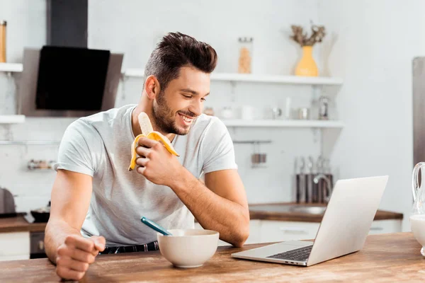 Uśmiechnięty Mężczyzna Młody Jedzenie Banana Patrząc Laptopie Kuchni — Zdjęcie stockowe