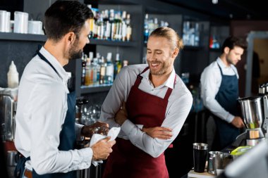 cheerful smiling barmen standing with arms crossed at workplace while coworker polishing glass clipart