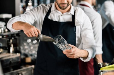 cropped view of  barman in apron putting ice in glass with ice shovel clipart
