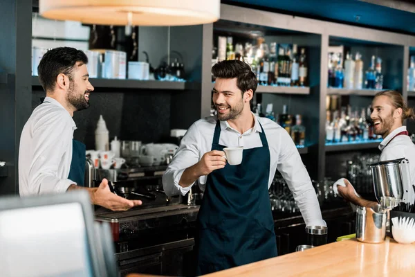 handsome cheerful barman drinking coffee and speaking with colleagues at workplace