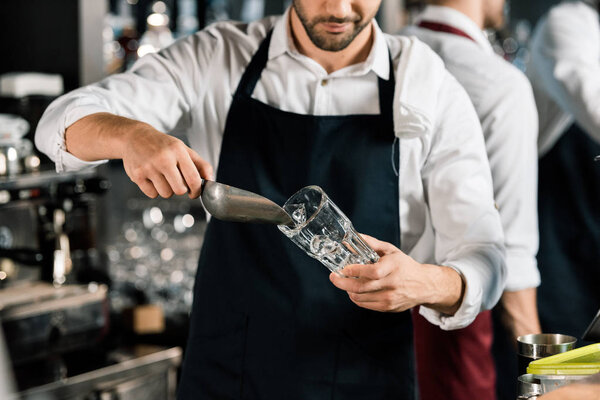 cropped view of  barman in apron putting ice in glass with ice shovel