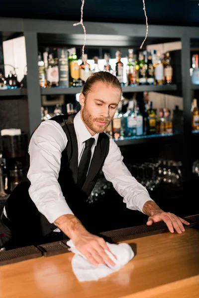 Handsome barman cleaning wooden bar counter with white napkin