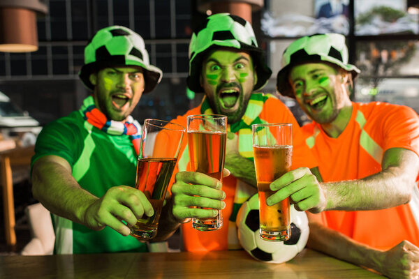 Футбольные болельщики держат бокалы с пивом и кричат в баре

