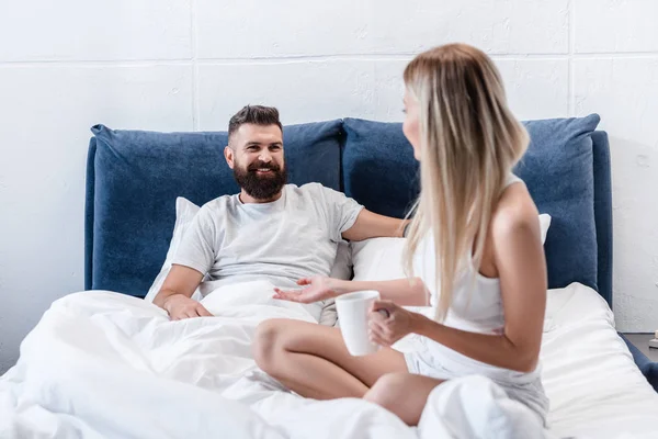 金发碧眼的女孩与咖啡杯说话与微笑的胡须男子躺在白色枕头在床上 — 免费的图库照片