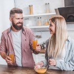 Joven mujer preparando el desayuno mientras alegre hombre sosteniendo vasos con jugo