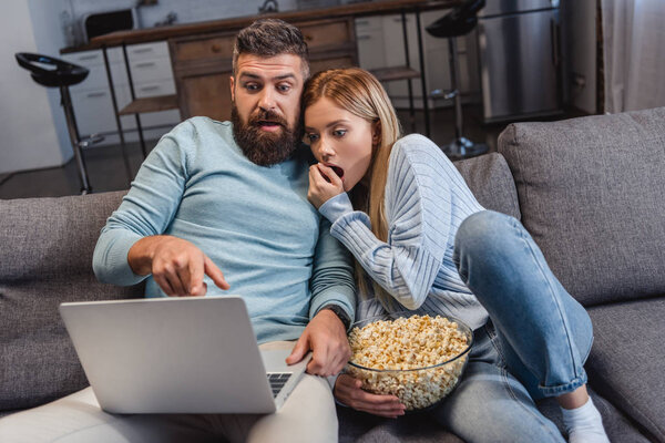 Муж и жена сидят на диване и смотрят страшный фильм на ноутбуке.
 
