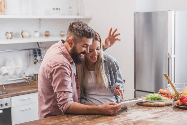 cheerful couple using digital tablet and gesturing at kitchen