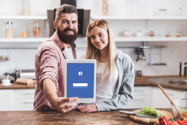 couple holding digital tablet with facebook app on screen at kitchen
