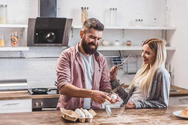 handsome bearded man smashing eggs at wooden table while woman holding balloon whisk