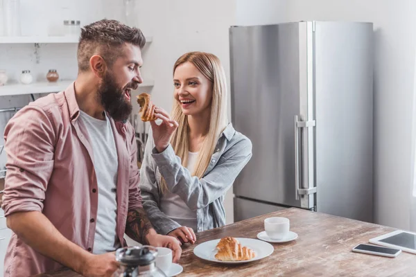 Cheerful Blonde Woman Feeding Bearded Boyfriend Croissant Kitchen — Stock Photo, Image