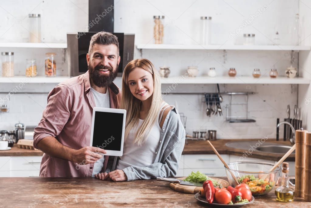 happy couple holding digital tablet with blank screen at wooden kitchen table