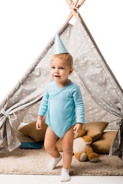 toddler boy in party hat standing in front of wigwam with pillows and teddy bear isolated on white clipart