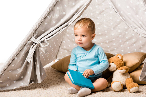toddler boy holding blue book and sitting in wigwam with pillows and teddy bear isolated on white