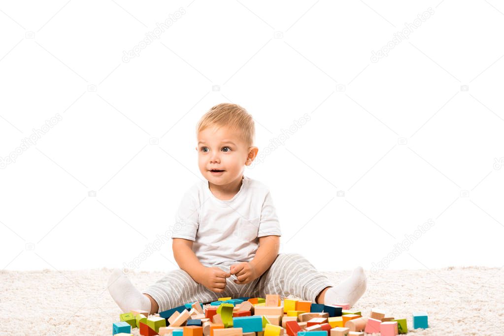 smiling toddler boy sitting on carpet and playing with multicolored wooden building blocks isolated on white
