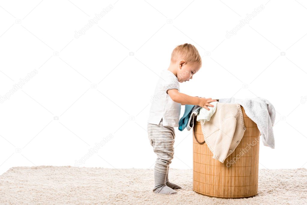 toddler boy standing in front of wicker laundry basket on carpet isolated on white