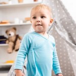 Cute toddler boy in blue bodysuit standing near grey wigwam in nursery room