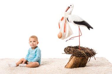 adorable toddler boy in blue bodysuit sitting on carpet near big decorative stork isolated on white clipart