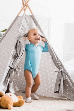 cute toddler boy in blue bodysuit smiling and standing in grey baby wigwam in nursery room clipart