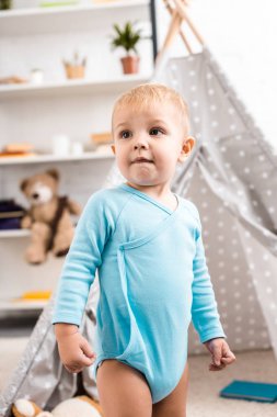 cute toddler boy in blue bodysuit standing near grey wigwam in nursery room clipart