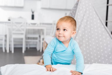 cute toddler boy standing near bed and smiling in apartment clipart