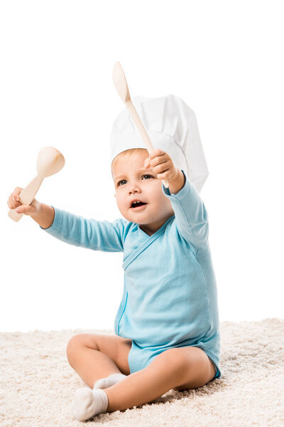 toddler boy in chefs hat sitting on carpet and holding two big wooden spoons isolated on white