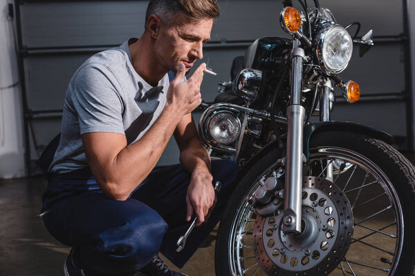 handsome adult mechanic smoking and holding wrench while fixing motorcycle in garage