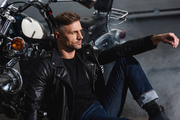 красивый байкер в кожаной куртке расслабляющий на мотоцикле в гараже
