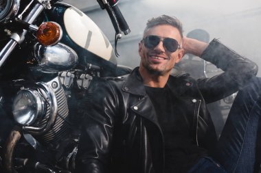 handsome man in sunglasses smiling sitting by motorcycle in garage clipart