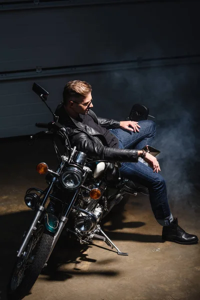 ガレージでのバイクに座っているサングラスでハンサムな男の側面図  — 無料ストックフォト
