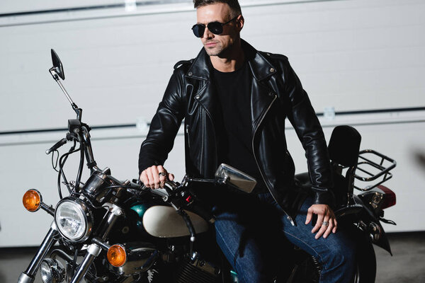 handsome classic man in black sunglasses and leather jacket standing by motorcycle in garage