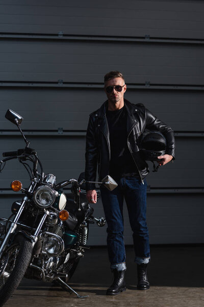 stylish biker in black sunglasses and leather jacket standing by motorbike and holding helmet