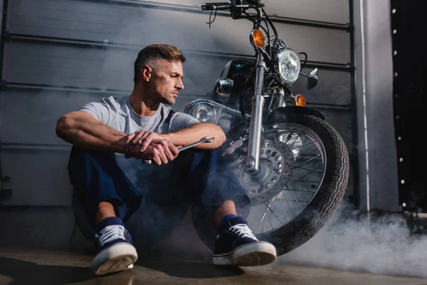Przystojny Mechanik Odwracając Trzymając Klucz Siedzi Pobliżu Motocykl Garażu — Zdjęcie stockowe