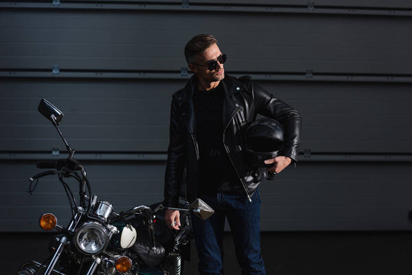 handsome biker in sunglasses holding helment and standing by motorcycle in garage