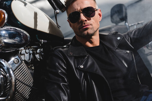 handsome classic guy in sunglasses sitting by motorcycle in garage