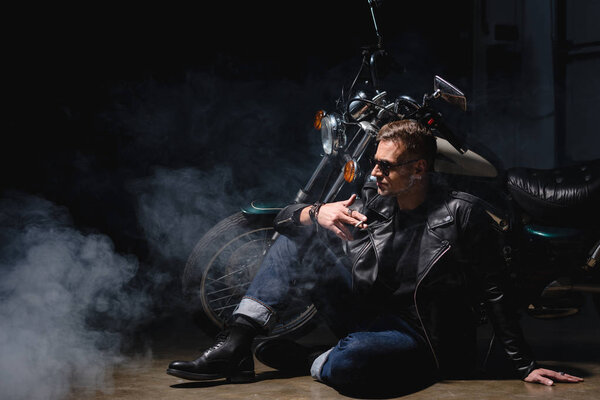 handsome biker in black sunglasses sitting on floor next to motorcycle in garage