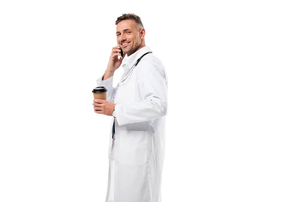 微笑的医生看着相机 拿着咖啡去和智能手机上的交谈孤立的白色 — 免费的图库照片