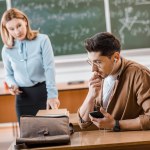 Estudante em óculos usando fones de ouvido e conversando no smartphone em sala de aula