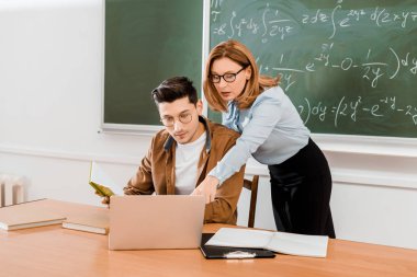 Female teacher pointing at laptop near student in class clipart