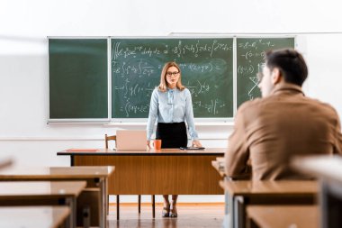 Sınıf denklemler ile kara tahta yakınındaki masasında duran kadın öğretim üyesi