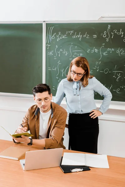 Młody Student Robiąc Notatki Patrząc Laptopie Pobliżu Nauczyciela Klasie — Darmowe zdjęcie stockowe