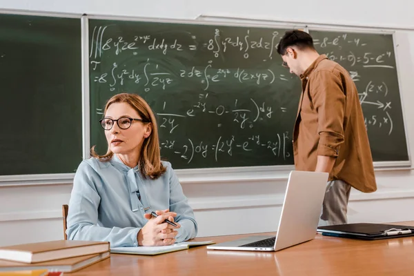 Nauczycielka Siedzi Przy Biurku Pobliżu Laptop Student Stojący Pobliżu Chalkboard — Zdjęcie stockowe